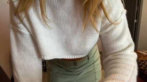 Media: A video of a young woman with blonde hair wearing a white, knitted crop top and high-waisted, light green pants, leaning slightly forward, in a cozy indoor setting.