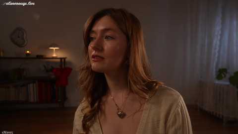 Media: Video of a fair-skinned woman with long, wavy auburn hair, wearing a beige top, standing in a dimly lit, cozy room with wooden floors and a shelf of books.