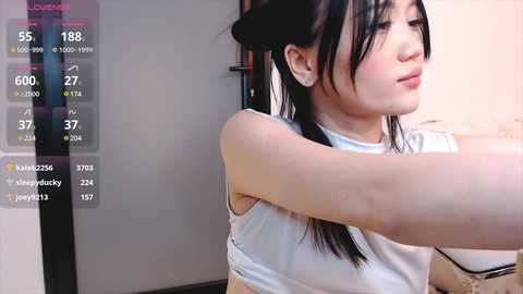 Media: A video of an East Asian woman with straight black hair, wearing a white sleeveless top, stretching her arm in a domestic setting. A virtual fitness app overlay displays her exercise stats.