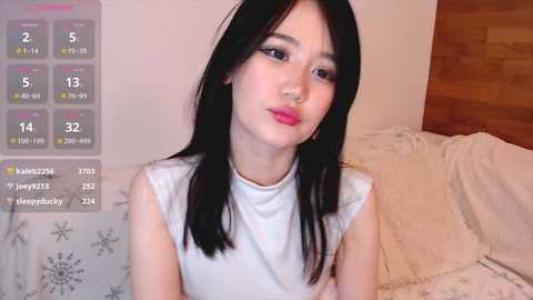 Media: Video of an East Asian woman with long black hair, light skin, and pink lipstick, wearing a sleeveless white top, in a cozy bedroom with a wooden headboard and a bed covered with a white duvet.