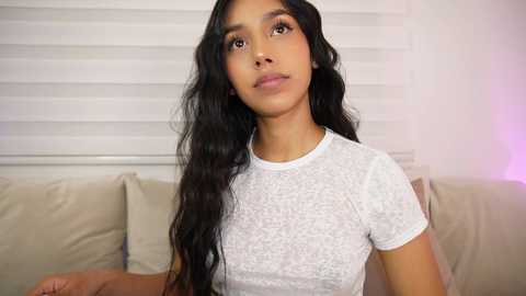 Video of a young South Asian woman with long black wavy hair, light brown skin, and dark eyes, wearing a white short-sleeved shirt. She sits on a beige couch against a white wall with horizontal blinds.