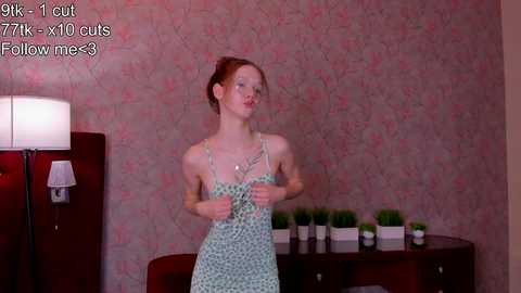 Media: Video of a slender, red-haired woman in a green polka-dot dress, standing in a room with pink floral wallpaper, dark furniture, and potted plants.