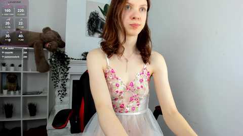 Media: Video of a young woman with fair skin and red hair, wearing a sheer floral dress, sitting in a modern living room with a white wall and a black and red gaming chair.