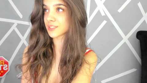 Media: Video of a young woman with long, wavy brown hair, light skin, wearing a red top, smiling against a gray geometric patterned wall.