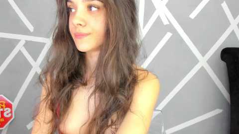 Media: Video of a topless young woman with long, wavy brown hair, light skin, and a slender physique. She has a confident expression and is positioned against a modern, geometric-patterned gray wall.