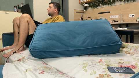 Media: Video of a man sitting on a floral-patterned couch with a blue pillow, wearing a mustard-yellow t-shirt and shorts, in a modern kitchen with wooden cabinets and a green wall.