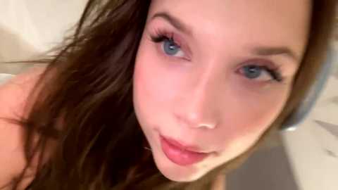 Close-up video of a young woman with light skin, blue eyes, and long brown hair, wearing pink lipstick, possibly in a bathroom.