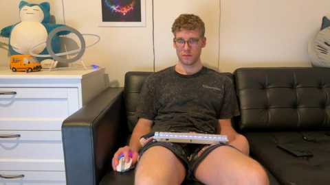 Media: Video of a young man with glasses and short brown hair, wearing a dark t-shirt and shorts, sitting on a black leather couch. He holds a Wii Remote and a game controller, surrounded by toy figures and a white dresser in a brightly lit room.