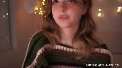 Media: A video of a young woman with fair skin and light brown hair, wearing a knitted sweater with horizontal stripes, gazing upwards with a neutral expression. Background features warm, soft lighting and bokeh effects.