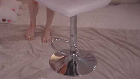 Media: A video of a woman's bare feet on a shiny chrome barstool base with a white cushioned seat, set against a beige carpeted background.