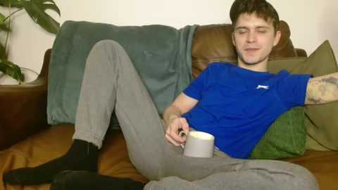 Media: Video of a young man with short brown hair, wearing a blue t-shirt and grey sweatpants, relaxing on a brown leather couch, holding a white mug.