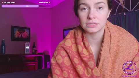 Media: Video of a woman with short dark hair wrapped in an orange polka-dot towel, sitting in a dimly lit room with purple lighting, framed art, and a dark wooden dresser.