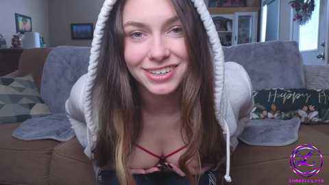 Media: Video of a young woman with long brown hair, wearing a gray hoodie, exposing a red bra, smiling on a beige sofa.