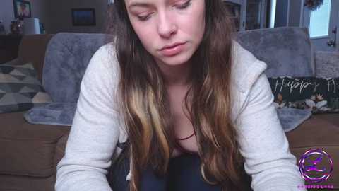 Media: A video of a young woman with long, wavy brown hair, wearing a gray hoodie and dark pants, leaning forward, sitting on a beige sofa in a cozy living room.