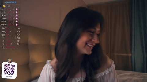 Media: Video of a smiling South Asian woman with long black hair, wearing a white off-shoulder top, standing in a dimly lit bedroom with a headboard and curtains.