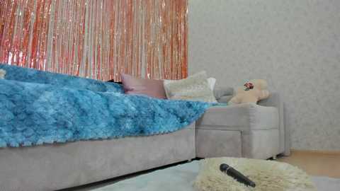 Media: Video of a cozy bedroom with a plush, blue faux fur throw on a grey couch. A teddy bear and a remote control lie on the floor. Orange tinsel streamers and a light grey patterned wallpaper adorn the background.