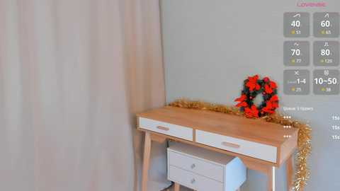 Media: Video of a wooden desk with white drawers, a red and black floral arrangement, a gold tinsel garland, and a white chair. The background features a light gray wall and beige curtains.