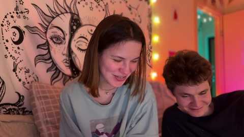 Media: Video of a young woman with shoulder-length brown hair, smiling, in a light blue shirt, sitting next to a young man with short brown hair, wearing a black shirt, in a cozy, dimly-lit room with a tapestry of suns and stars on the wall.