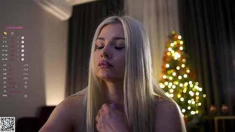 Media: Video of a fair-skinned, blonde woman with long hair, wearing a white top, standing indoors by a lit Christmas tree.