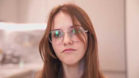 Media: A video of a young woman with fair skin, straight auburn hair, and round, metal-framed glasses, standing in a blurred bathroom setting.