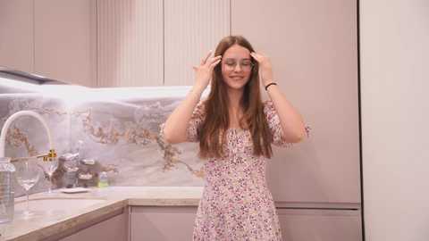 Media: Video of a young woman with long, straight brown hair, wearing a floral dress, adjusting her glasses in a modern, light-colored kitchen with a marble backsplash.