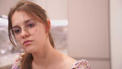 Media: Video of a young Caucasian woman with fair skin and brown hair, wearing round glasses and a floral-patterned off-shoulder top, standing indoors with blurred background.