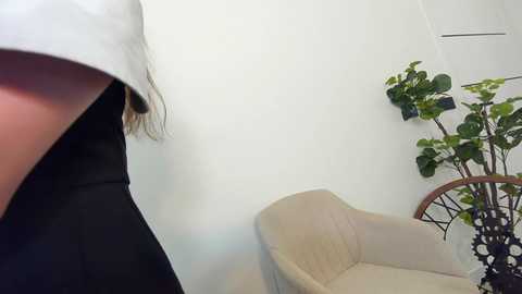 Media: Video of a woman with long brown hair wearing a white shirt and black jacket, standing in a minimalist room with a beige armchair, green potted plant, and white walls.