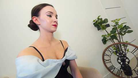 Media: Video of a fair-skinned woman with dark hair in a bun, wearing a light blue off-shoulder top and black bra, standing against a plain white wall with a green plant and antique clock in the background.