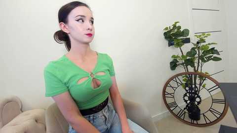 Media: Video of a young woman with pale skin and dark hair tied back, wearing a green crop top with cutouts and light-wash jeans, sitting on a beige couch in a minimalist room with a large clock and potted plant.