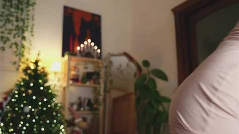 Media: Video of a warmly lit living room with a decorated Christmas tree, a lit candle centerpiece, a green plant, and a person in a beige shirt seen from the back.