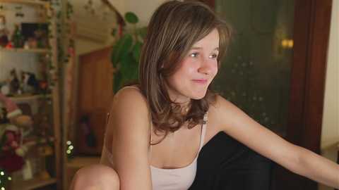 Media: Video of a young Caucasian woman with light brown hair, wearing a white spaghetti strap top, sitting on a black couch, indoors, looking thoughtful.