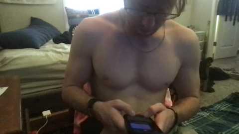 Media: Video of a shirtless man with glasses and a beard, wearing a black wristband, holding a phone, standing in a cluttered bedroom with a bed, carpet, and various objects.