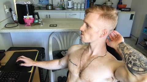 Media: Video of a shirtless, muscular, light-skinned man with short, blond hair, tattoos on his left arm, sitting at a computer in a modern kitchen, holding his ear with his right hand.