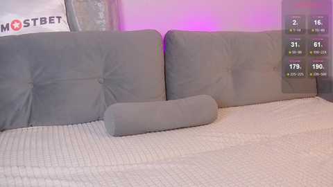 Media: A video of a modern, minimalist living room featuring a beige sectional sofa with gray cushions and a matching throw pillow. The background includes a purple wall and a visible \"MOSTBET\" banner.