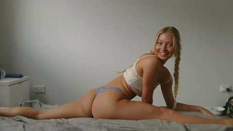 Media: Video of a young Caucasian woman with blonde braided hair, smiling, in a white sports bra and blue thong, performing a side split on a gray bedspread in a minimalist bedroom.