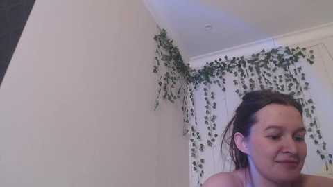 Media: Video of a young woman with light skin and brown hair, smiling, standing in a room with white walls and a green ivy-covered window.