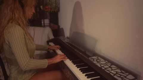 A video of a woman with long, wavy blonde hair, wearing a green sweater, playing a black and white keyboard in a dimly lit room with a white wall and shadowed corner.