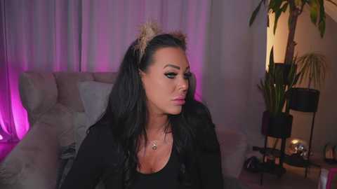 Media: Video of a woman with long black hair, wearing a black top, sitting in a dimly lit room with purple lighting, green plants, and a black lamp.