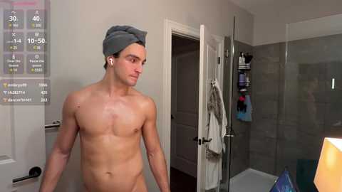 Media: Video of a shirtless, light-skinned man with a gray towel on his head, standing in a modern bathroom with a digital thermometer and a shower with towels hanging.