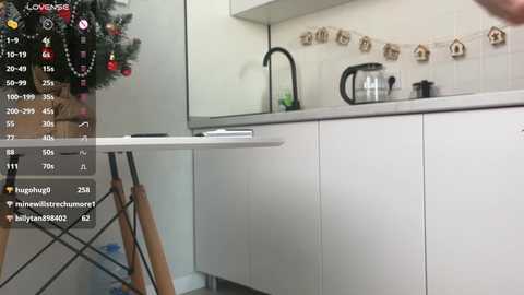 Video of a modern kitchen with white cabinets, a stainless steel sink, a glass kettle, and a Christmas tree adorned with red and gold decorations.