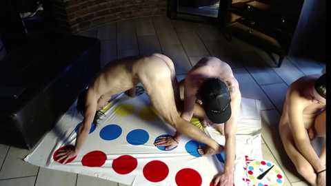 Media: Video of three nude men playing Twister on a tiled floor, one man blindfolded, another bent over, and a third sitting.