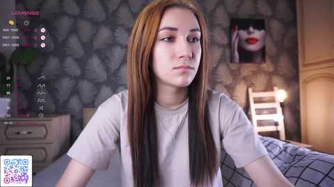 Media: Video of a young woman with long, straight, brown hair, wearing a beige t-shirt, sitting on a bed in a dimly lit room with a patterned wallpaper and a nightstand.