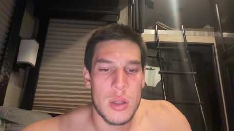 Media: Video of a shirtless young man with short dark hair, slightly flushed face, and closed eyes, standing indoors near a closed garage door and a bunk bed.