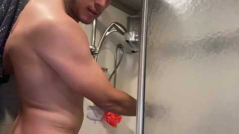 Media: Video of a shirtless, muscular man with tanned skin, wet hair, and a slight smile, showering in a steamy bathroom with a metallic faucet and red loofah.