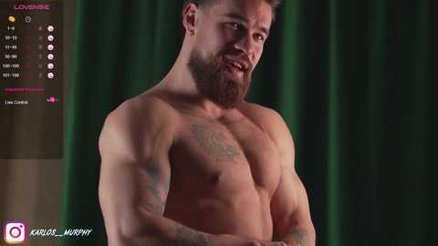 Video of a muscular, shirtless man with a beard, tattoos, and a confident expression, standing against dark green curtains.