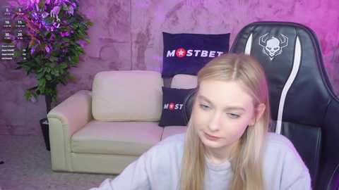 Media: Video of a young, light-skinned, blonde woman in a gray sweater, seated in a black gaming chair with a skull emblem, in a room with a cream couch, purple wall, and plant.