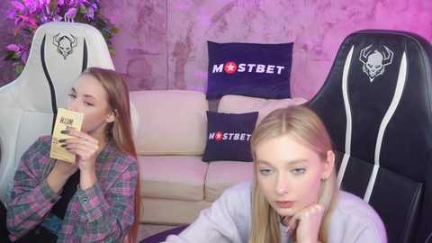 Media: Video of two young women with long hair, one in a checkered shirt, the other in a gray sweater, seated on gaming chairs in a dimly lit room with a white sofa and \"MSTBET\" pillows.