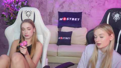 Video of two young women, one blonde and one brunette, sitting in white gaming chairs, holding pink vibrators, in a room with a cream sofa, dark cushions, and a purple wall.