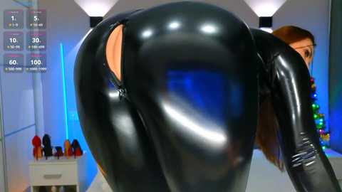 Media: Video of a woman in a tight, shiny black latex bodysuit, emphasizing her large, round buttocks. The background shows a modern, brightly lit room with blue lighting and a white dresser with red objects.