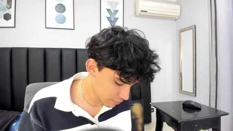 Media: Video of a young Hispanic man with curly black hair, wearing a white and navy polo shirt, sitting in a modern room with black leather furniture, white walls, and minimalist decor.